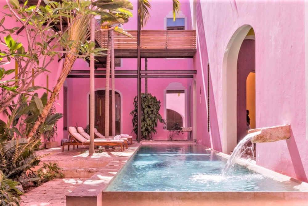 Rosas y Xocolate hotel in Merida, Mexico. A pool is fed by a small fountain sprouting from the rose pink facade of the hotel in a partially shaded courtyard.